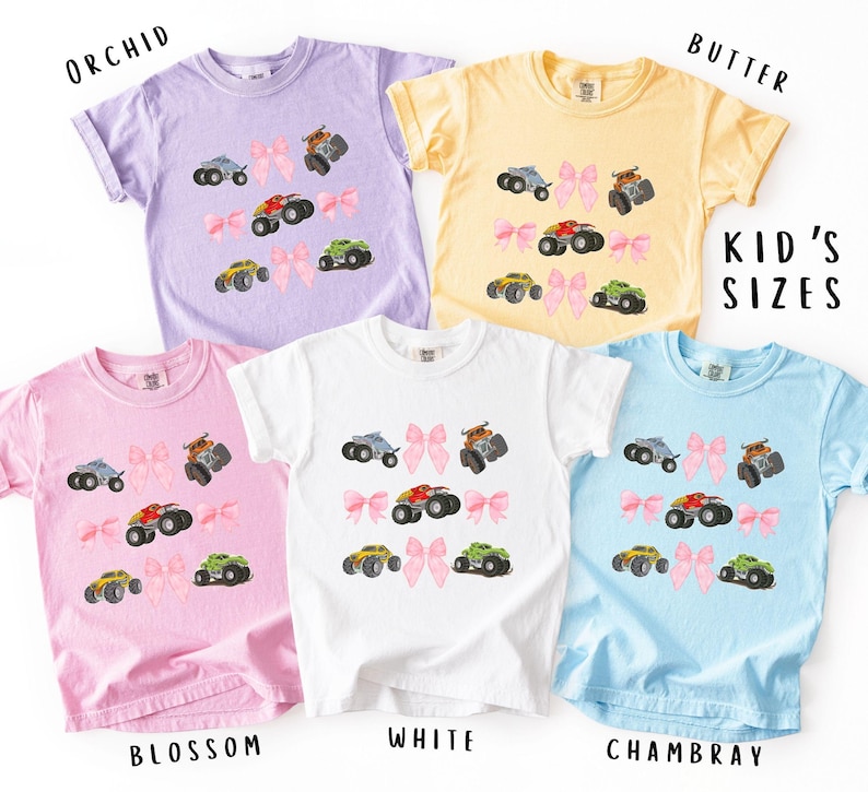 K&ouml;nnte beinhalten: Eine Auswahl an Kinder-T-Shirts in den Farben Orchidee, Butter, Blossom, Wei&szlig; und Chambray. Jedes Shirt zeigt ein Monster-Truck-Design mit einer rosa Schleife. Der Text "KID'S SIZES" ist ebenfalls sichtbar.