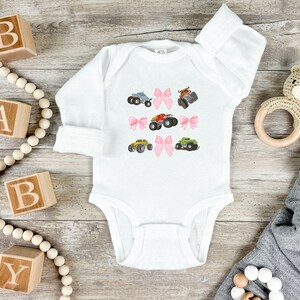 May include: White baby onesie with long sleeves, decorated with colorful monster truck illustrations and pink bows. The onesie is made of soft material and is suitable for infants. The design includes trucks in blue, brown, red, yellow, and green.