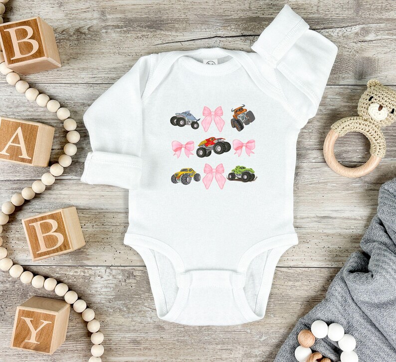 May include: White baby onesie with long sleeves, decorated with colorful monster truck illustrations and pink bows. The onesie is made of soft material and is suitable for infants. The design includes trucks in blue, brown, red, yellow, and green.
