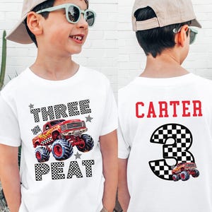 Pictured is a boy wearing a light colored t-shirt which says THREE PEAT in black ans white checkered letters along with a big red monster truck graphic and black and white checkered stars and flag. On the back of the shirt is custom name CARTER in red and a black and white checkered number 3