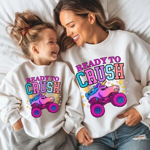 Pu&ograve; includere: Felpe bianche abbinate con la scritta "READY TO CRUSH" e un disegno di monster truck unicorno. Una madre e suo figlio sorridenti le indossano.