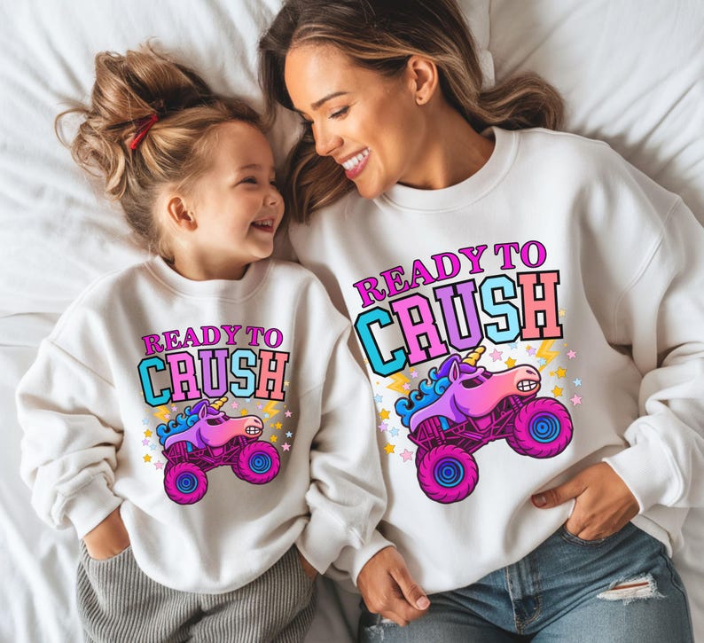 Pu&ograve; includere: Felpe bianche abbinate con la scritta "READY TO CRUSH" e un disegno di monster truck unicorno. Una madre e suo figlio sorridenti le indossano.