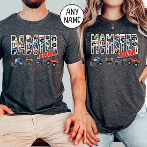 May include: Two gray t-shirts with a monster truck theme. The shirt on the left says "DADSTER JAM" and the shirt on the right says "MOMSTER JAM". Both shirts feature a line of monster trucks with colorful designs.