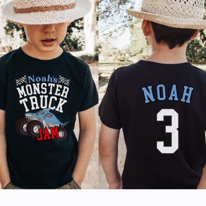 以下が含まれることがあります： サメと「Noah's Monster Truck Jam」の文字が入った、モンスター トラックのグラフィックが特徴の黒いTシャツ。Tシャツの背面には、名前「Noah」と数字「3」が青と白で表示されています。