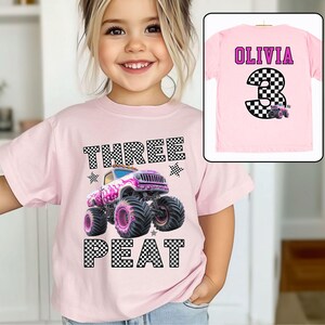 Pictured is a young girl wearing a pink shirt which says THREE PEAT in black and white checkered letters along with a big graphic of a pink monster truck and checked stars and flag. On the back of this tee is custom name OLIVIA in purple and custom number (Age) 3 which is also checkered