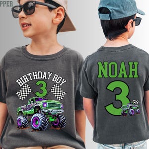 Pode incluir: Camiseta cinza escuro com um gr&aacute;fico de monster truck verde e roxo. A frente da camisa diz "BIRTHDAY BOY 3" com bandeiras quadriculadas. A parte de tr&aacute;s da camisa diz "NOAH 3".