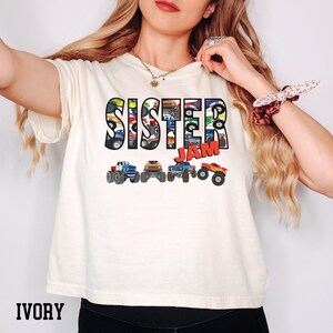 K&ouml;nnte beinhalten: Ein T-Shirt in Elfenbein mit den Worten "Sister Jam" in schwarzen und roten Buchstaben. Die Buchstaben bestehen aus Bildern von Monstertrucks.