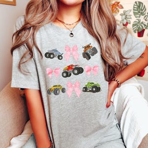Op de afbeelding: Een lichtgrijs t-shirt met kleurrijke monstertruckafbeeldingen en roze strikken. De trucks zijn in verschillende kleuren, waaronder blauw, oranje, rood, geel en groen. Het ontwerp is speels en geschikt voor kinderen.