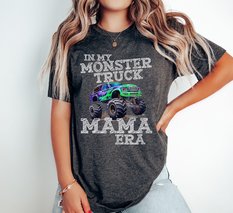 A woman wearing a dark gray t-shirt with a graphic of a green, purple, and black monster truck with the text "In My Monster Truck Mama Era" in white chalk text