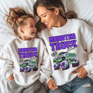 May include: A white sweatshirt with the text "Monster Truck Jam" in purple letters and a green monster truck graphic. A matching smaller sweatshirt with the same design is worn by a child.