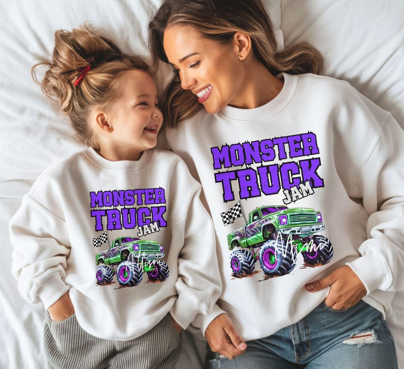 May include: A white sweatshirt with the text "Monster Truck Jam" in purple letters and a green monster truck graphic. A matching smaller sweatshirt with the same design is worn by a child.