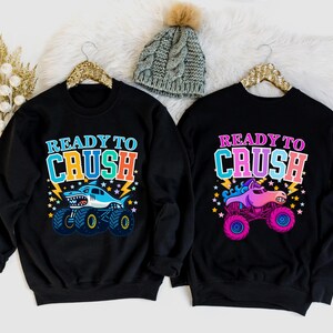 Pu&ograve; includere: Due felpe nere con la scritta "READY TO CRUSH". Una presenta un monster truck a forma di squalo, l'altra un monster truck a forma di unicorno. Entrambe hanno disegni colorati con fulmini e stelle. &Egrave; visibile anche un berretto di lana grigio con un pompon.