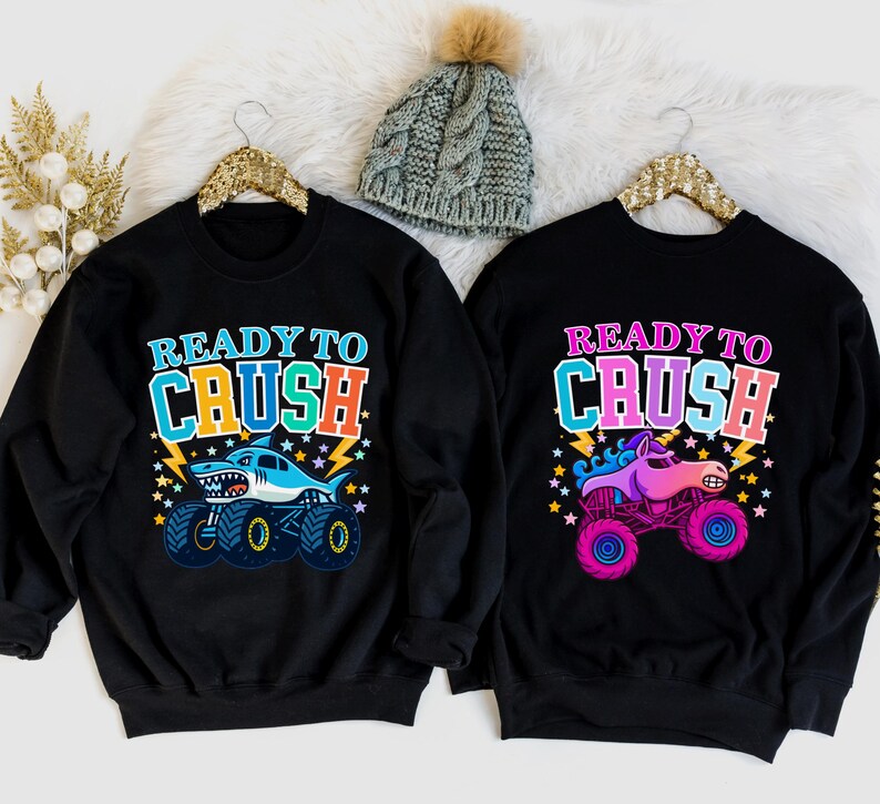 Pu&ograve; includere: Due felpe nere con la scritta "READY TO CRUSH". Una presenta un monster truck a forma di squalo, l'altra un monster truck a forma di unicorno. Entrambe hanno disegni colorati con fulmini e stelle. &Egrave; visibile anche un berretto di lana grigio con un pompon.