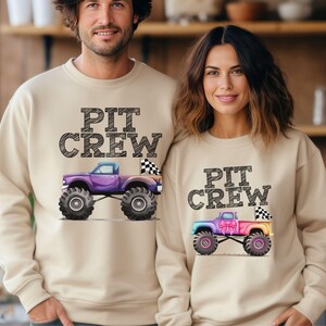 Puede incluir: Sudaderas beige con el texto "PIT CREW" sobre un gr&aacute;fico de monster truck. Un cami&oacute;n es morado, el otro es de color arco&iacute;ris. Ambos tienen neum&aacute;ticos grandes y banderas a cuadros. Las sudaderas son usadas por dos personas.
