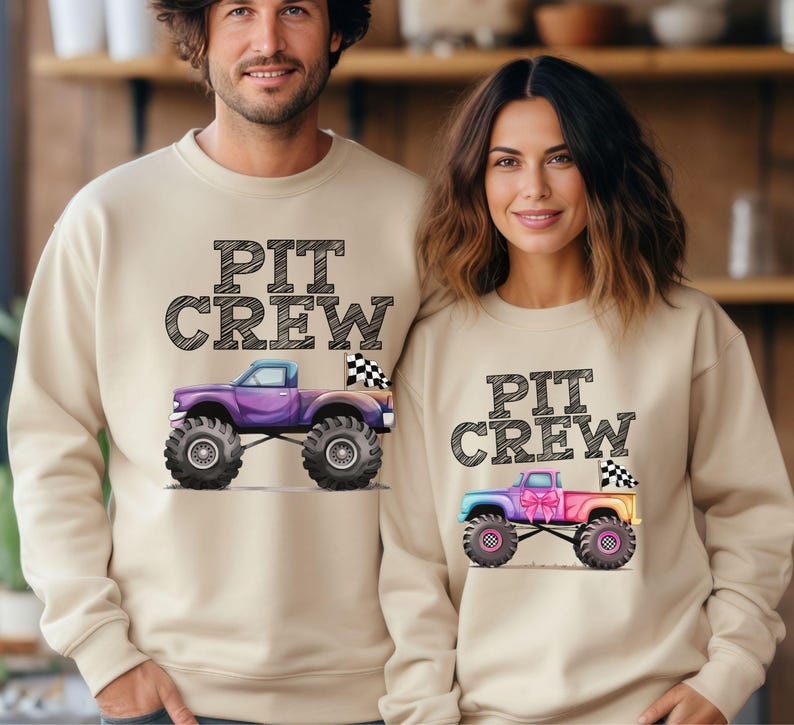 Puede incluir: Sudaderas beige con el texto "PIT CREW" sobre un gr&aacute;fico de monster truck. Un cami&oacute;n es morado, el otro es de color arco&iacute;ris. Ambos tienen neum&aacute;ticos grandes y banderas a cuadros. Las sudaderas son usadas por dos personas.