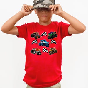 May include: A red t-shirt featuring colorful monster truck graphics and checkered flags. The shirt has a crew neck and short sleeves. The design includes various monster trucks in orange, green, blue, and red.