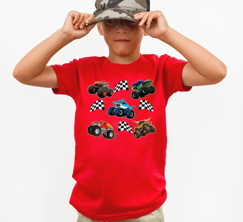 May include: A red t-shirt featuring colorful monster truck graphics and checkered flags. The shirt has a crew neck and short sleeves. The design includes various monster trucks in orange, green, blue, and red.