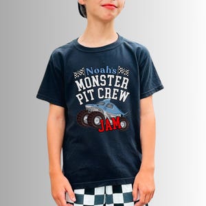 May include: Navy blue t-shirt with the text "Noah's Monster Pit Crew JAM" and a monster truck graphic. The graphic features a blue shark-shaped truck with large wheels. The shirt is worn by a person.