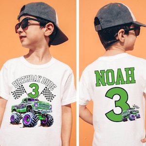 Pictured is a young boy wearing a natural colored comfort colors t-shirt with a front back print design theme. the front says BIRTHDAY DUDE and the number 3 with a purple and green Hot cars wheels monster truck theme and 2 checkered flags. the back has custom name NOAH and a big number 3 in green and a small version of the monster truck