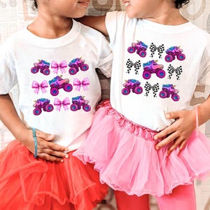 May include: Two white t-shirts with a repeating pattern of a monster truck with a unicorn horn and pink bows. The girls are wearing pink and red tutus.