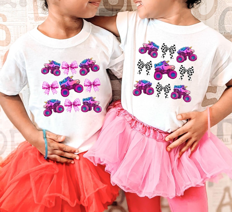May include: Two white t-shirts with a repeating pattern of a monster truck with a unicorn horn and pink bows. The girls are wearing pink and red tutus.