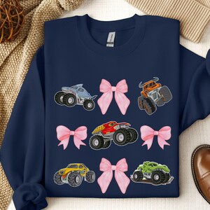 May include: Navy blue sweatshirt featuring colorful monster truck graphics and pink bows. The trucks are in various colors, including blue, orange, red, yellow, and green. A fun and playful design.