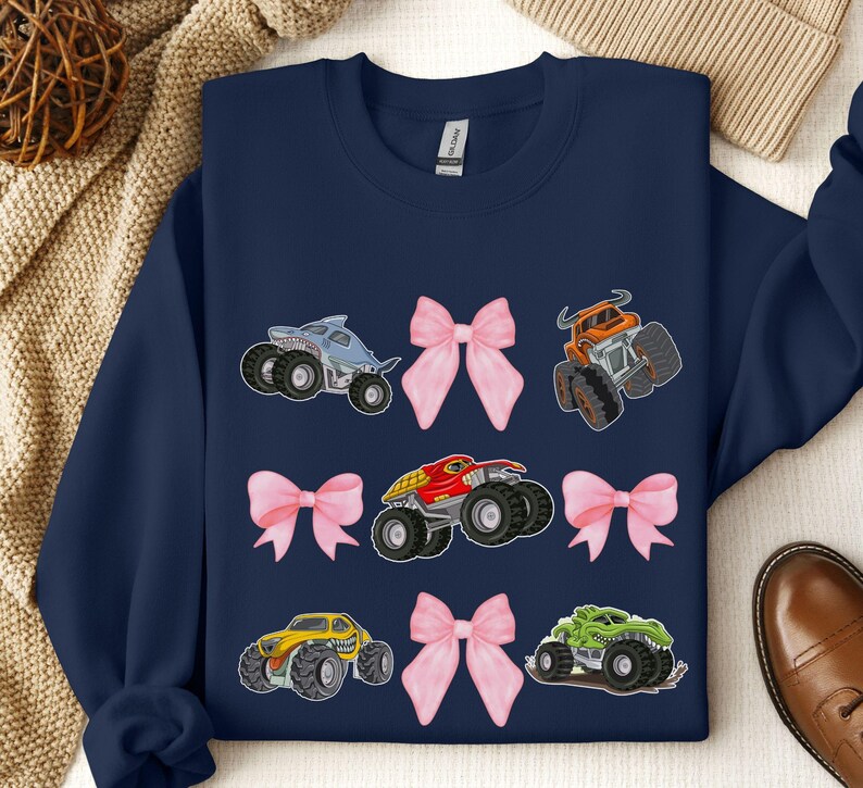 May include: Navy blue sweatshirt featuring colorful monster truck graphics and pink bows. The trucks are in various colors, including blue, orange, red, yellow, and green. A fun and playful design.