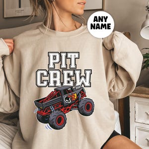 Pictured is a woman wearing a Beige sweatshirt with the text "PIT CREW" in a white and black big block font. Below the text is a graphic of a red and black monster truck with a skull and flames design. The sweatshirt has long sleeves.