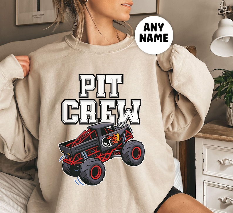 Pictured is a woman wearing a Beige sweatshirt with the text "PIT CREW" in a white and black big block font. Below the text is a graphic of a red and black monster truck with a skull and flames design. The sweatshirt has long sleeves.