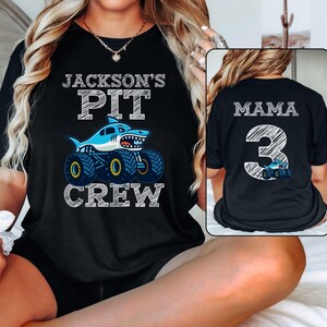 Op de afbeelding: Zwart T-shirt met de tekst "JACKSON'S PIT CREW" boven een cartoon haaien monstertruck. De achterkant van het shirt zegt "MAMA 3" met een bijpassende monstertruck afbeelding.
