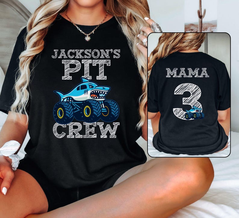 Op de afbeelding: Zwart T-shirt met de tekst "JACKSON'S PIT CREW" boven een cartoon haaien monstertruck. De achterkant van het shirt zegt "MAMA 3" met een bijpassende monstertruck afbeelding.