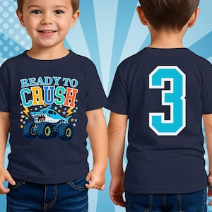 Pictured is a young boy wearing a navy blue t-shirt with a shark monster truck graphic and the text "READY TO CRUSH" with stars and lightning in the background. The back of the shirt features a large number "3" in turquoise with a white outline. The shirt is worn by a young child. No personal name for privacy.