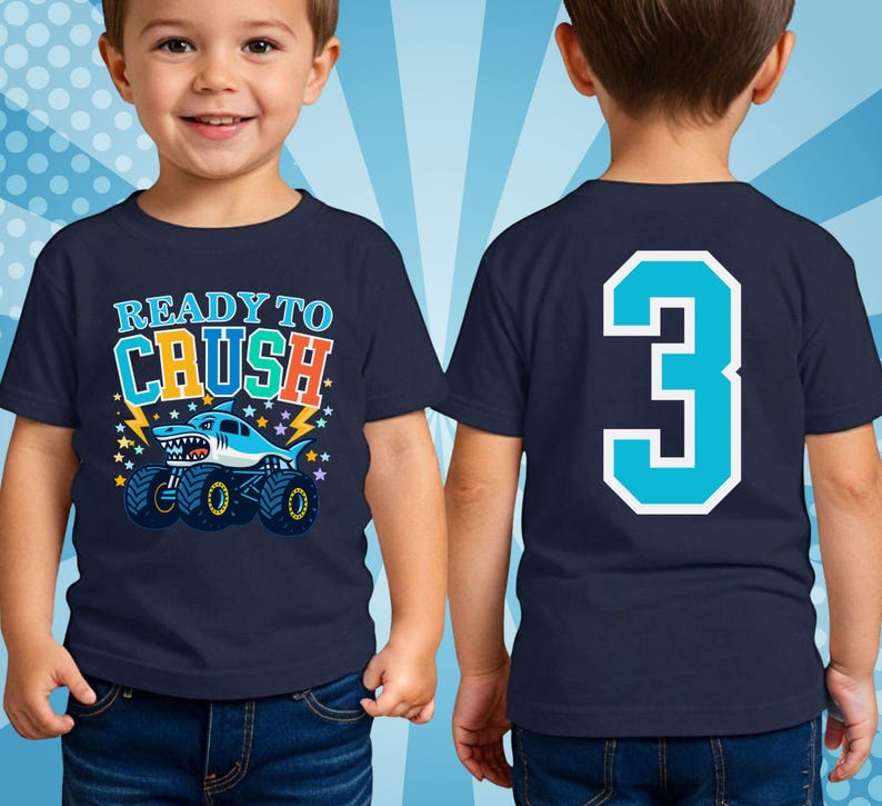 Pictured is a young boy wearing a navy blue t-shirt with a shark monster truck graphic and the text "READY TO CRUSH" with stars and lightning in the background. The back of the shirt features a large number "3" in turquoise with a white outline. The shirt is worn by a young child. No personal name for privacy.