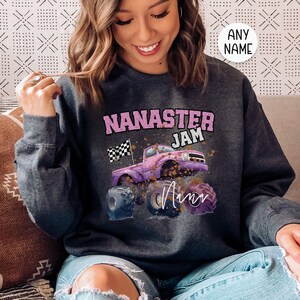 Pu&ograve; includere: Una felpa grigia scura con una grafica rosa e bianca di un monster truck con il testo "Nana's Jam" e la parola "Ann" scritta in corsivo.
