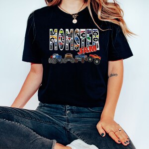 May include: A black t-shirt with the words "Monster Jam" printed in white with a colorful monster truck design. The shirt is being worn by a person wearing blue jeans.