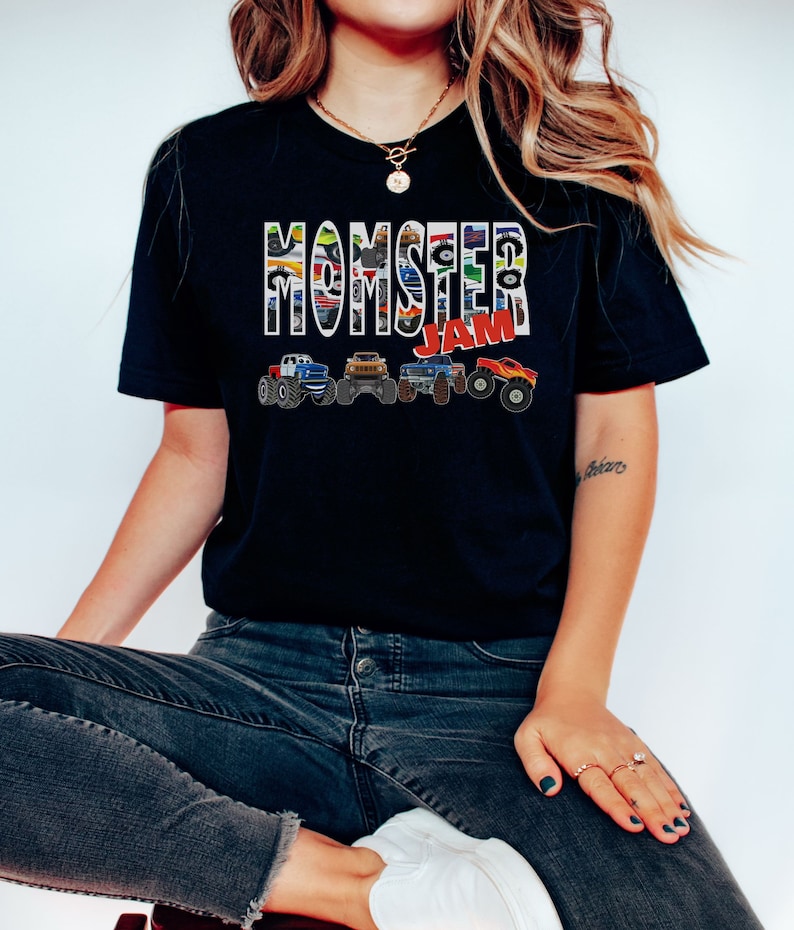 May include: A black t-shirt with the words "Monster Jam" printed in white with a colorful monster truck design. The shirt is being worn by a person wearing blue jeans.