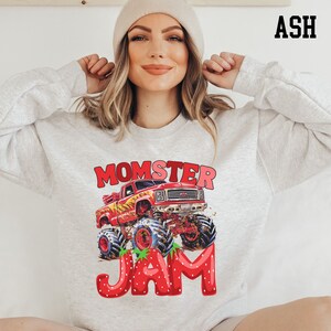 May include: Ash-colored sweatshirt featuring a red monster truck graphic with the text "MOMSTER JAM." The truck has large tires and is decorated with strawberries. The text "ASH" is printed in black in the upper right corner.