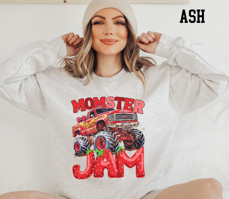 May include: Ash-colored sweatshirt featuring a red monster truck graphic with the text "MOMSTER JAM." The truck has large tires and is decorated with strawberries. The text "ASH" is printed in black in the upper right corner.