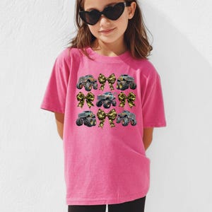 May include: A pink t-shirt featuring a design of monster trucks and bows. The monster trucks are in various colors, and the bows have a camouflage pattern. The shirt is worn by a young person wearing heart-shaped sunglasses.