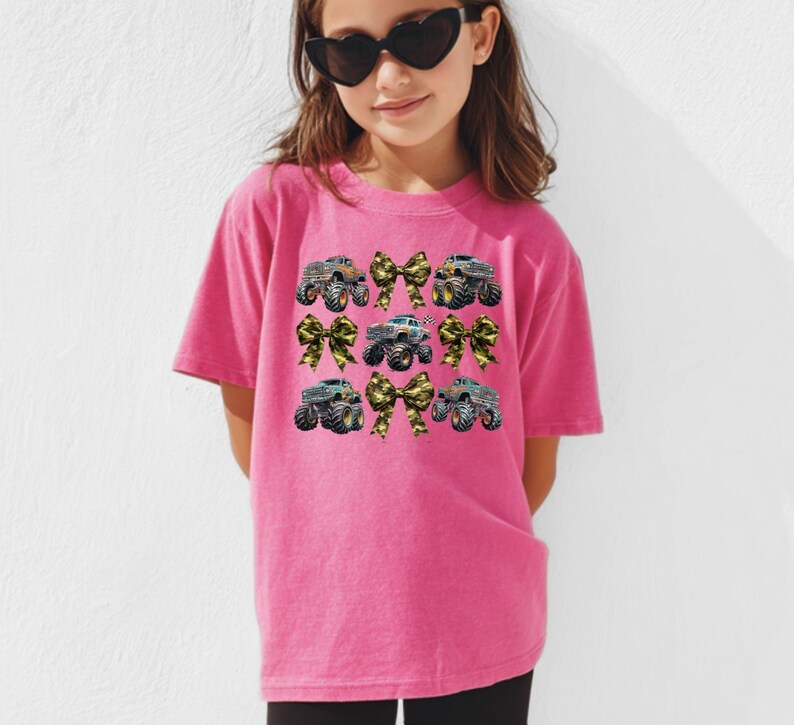May include: A pink t-shirt featuring a design of monster trucks and bows. The monster trucks are in various colors, and the bows have a camouflage pattern. The shirt is worn by a young person wearing heart-shaped sunglasses.