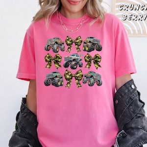 May include: A pink t-shirt featuring a design of monster trucks and camouflage bows. The text "CRUNCH BERRY" is printed in a script font at the top right. The shirt has short sleeves and a crew neck.