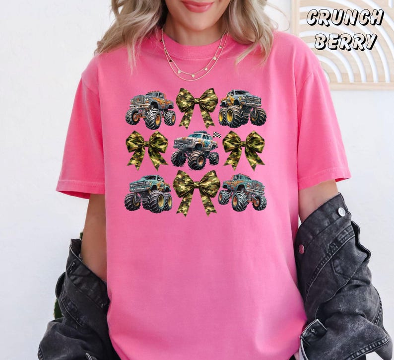 May include: A pink t-shirt featuring a design of monster trucks and camouflage bows. The text "CRUNCH BERRY" is printed in a script font at the top right. The shirt has short sleeves and a crew neck.