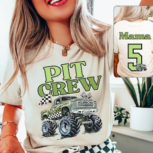Op de afbeelding: Beige T-shirt met een groene en zwarte monstertruckafbeelding en de woorden "PIT CREW" in het groen. De achterkant van de shirt heeft het woord "Mama", het nummer "5" en een monstertruckafbeelding. Het shirt heeft een ronde hals.