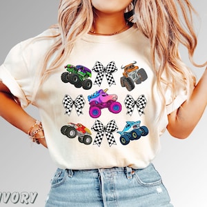 May include: Ivory-colored t-shirt featuring colorful monster truck graphics and checkered bows. The trucks include a green and purple truck, an orange truck, a unicorn truck, a red truck, and a blue shark truck. The word "IVORY" is printed at the bottom.