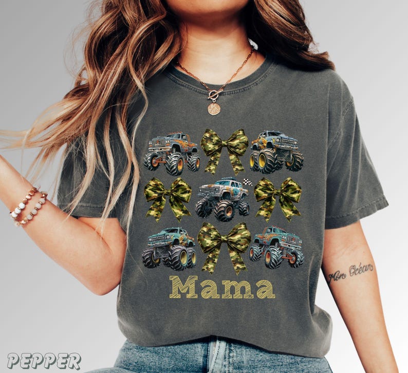Pictured is a woman wearing a dark gray t-shirt featuring a design of monster trucks and camouflage bows. The word "Mama" is printed in gold block letters. The shirt has a crew neck and short sleeves.