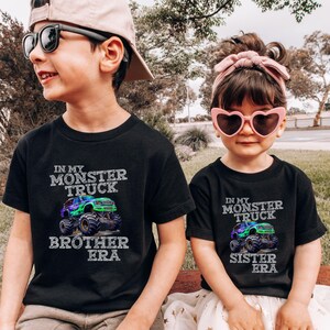 May include: Two children in black t-shirts featuring a monster truck design. The shirts display the text "In My Monster Truck Brother Era" and "In My Monster Truck Sister Era". The boy wears sunglasses and a cap, and the girl wears heart-shaped sunglasses and a pink headband.