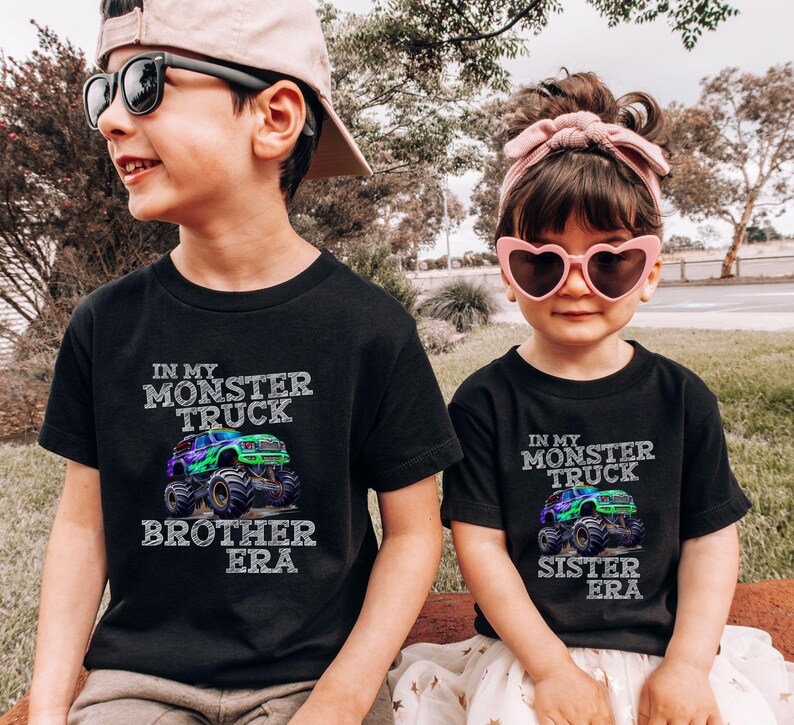 May include: Two children in black t-shirts featuring a monster truck design. The shirts display the text "In My Monster Truck Brother Era" and "In My Monster Truck Sister Era". The boy wears sunglasses and a cap, and the girl wears heart-shaped sunglasses and a pink headband.