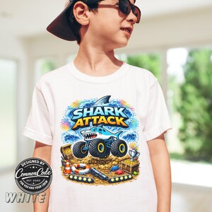 Puede incluir: Camiseta blanca con un gr&aacute;fico colorido de "Shark Attack" que presenta un cami&oacute;n monstruo con tem&aacute;tica de tiburones, cofres del tesoro y un salvavidas. La camiseta tambi&eacute;n tiene el texto "DESIGNED BY Common Code EST. 2020 FOR YOU AND YOUR CREW WHITE".