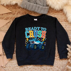 Pu&ograve; includere: Felpa nera con la scritta "READY TO CRUSH" in blu, arancione e verde. Un cartone animato di uno squalo su un monster truck &egrave; raffigurato sotto il testo. Lo squalo &egrave; azzurro con la pancia bianca e la bocca spalancata.
