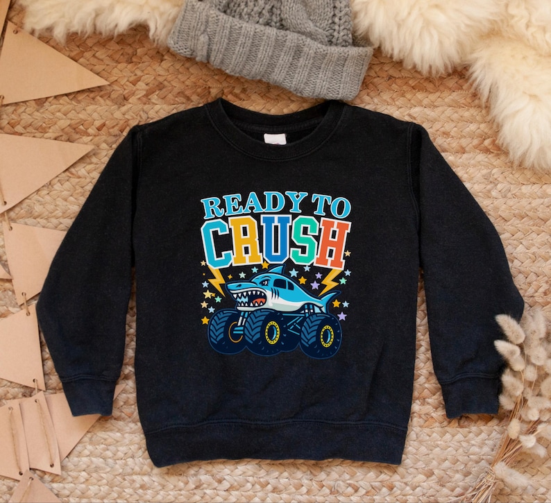 Pu&ograve; includere: Felpa nera con la scritta "READY TO CRUSH" in blu, arancione e verde. Un cartone animato di uno squalo su un monster truck &egrave; raffigurato sotto il testo. Lo squalo &egrave; azzurro con la pancia bianca e la bocca spalancata.